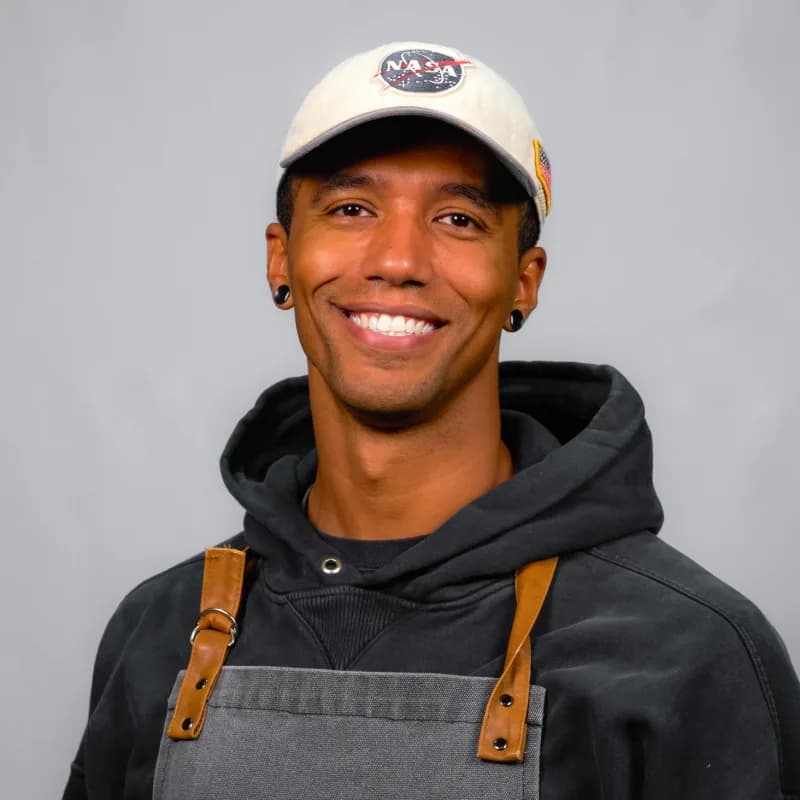 Milton Perry, smiling in a NASA cap, dark hoodie, and work apron
